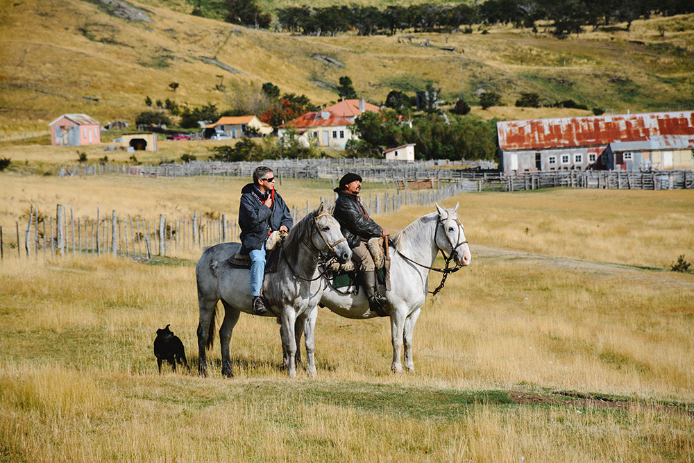 Two riders on horseback in front of a rural estate in Chilean Patagonia, with a landscape of fields and hills