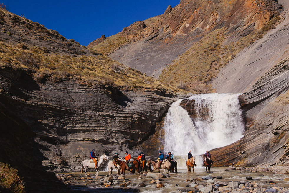 Group of riders on horseback in front of a waterfall in a rocky canyon in Chilean Patagonia