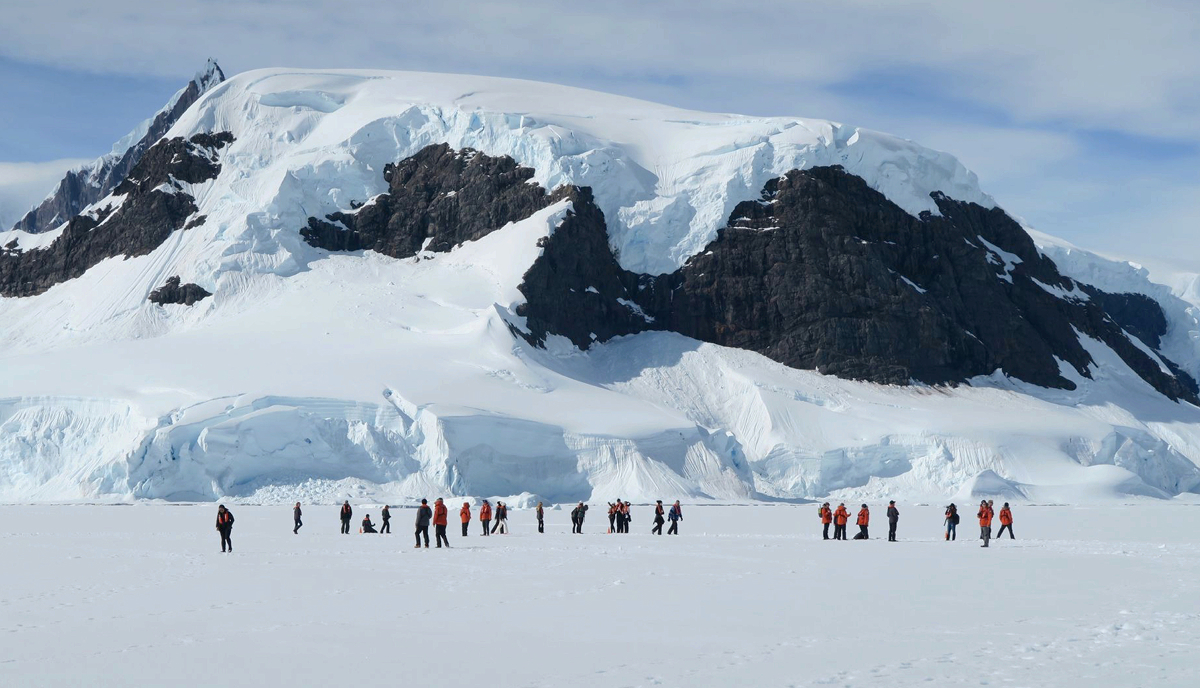 Group of people with orange jackets walking on the ice in front of a huge glacier in Antarctica.