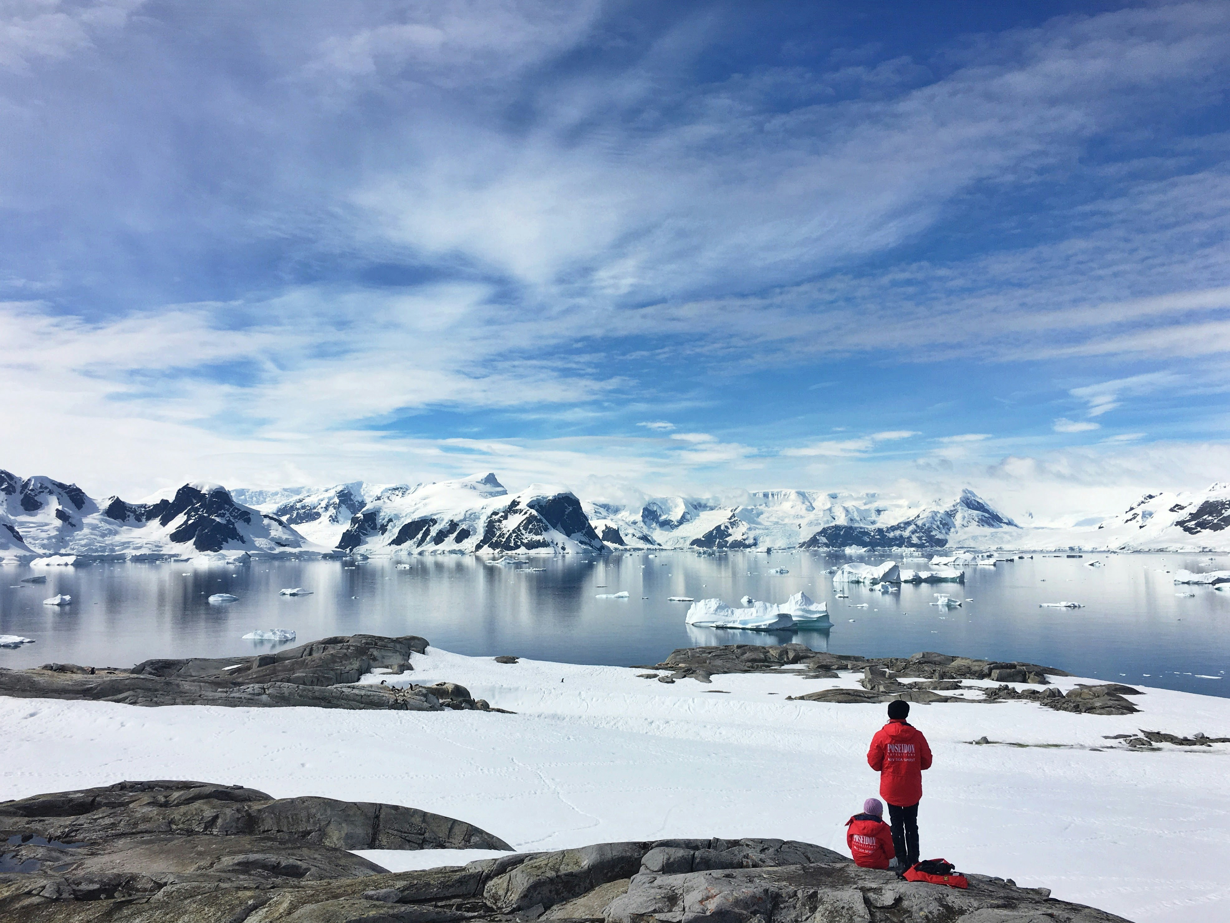 Two people in red jackets contemplating an Antarctic landscape of snow-covered mountains, floating icebergs, and a partially clear sky, from a rocky area.