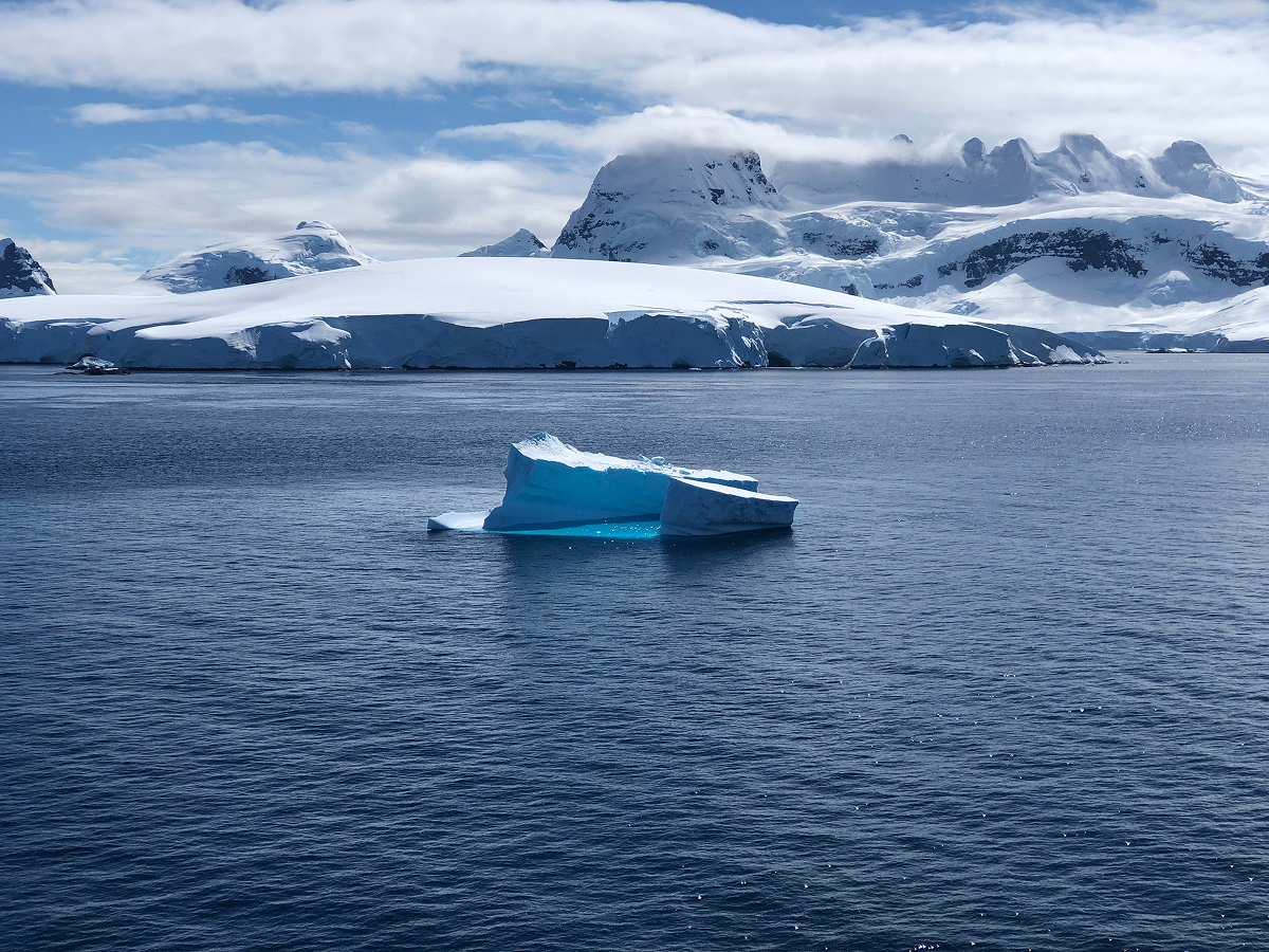Iceberg floating in the Antarctic Ocean with snow-covered mountains in the background and a partially cloudy sky.