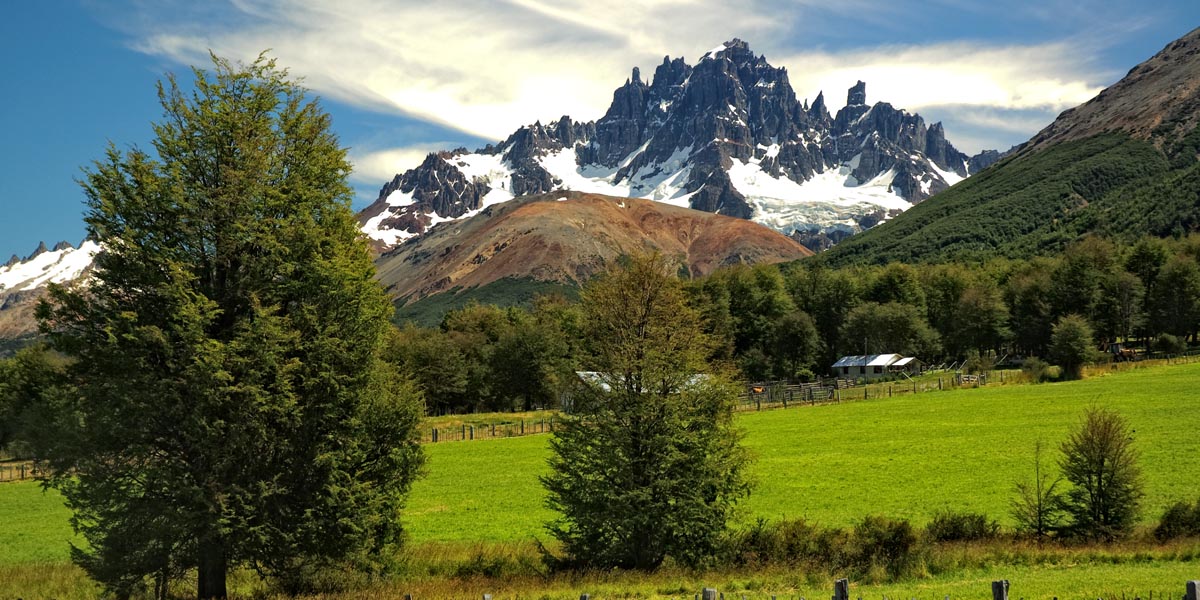 Panoramic view of Cerro Castillo National Park in Chilean Patagonia, with green meadows, native forests, and the imposing snow-covered rocky towers of Cerro Castillo in the background.