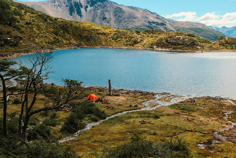 Campsite next to a turquoise lake in the Dientes de Navarino Circuit, surrounded by mountains and typical vegetation of Chilean Patagonia.