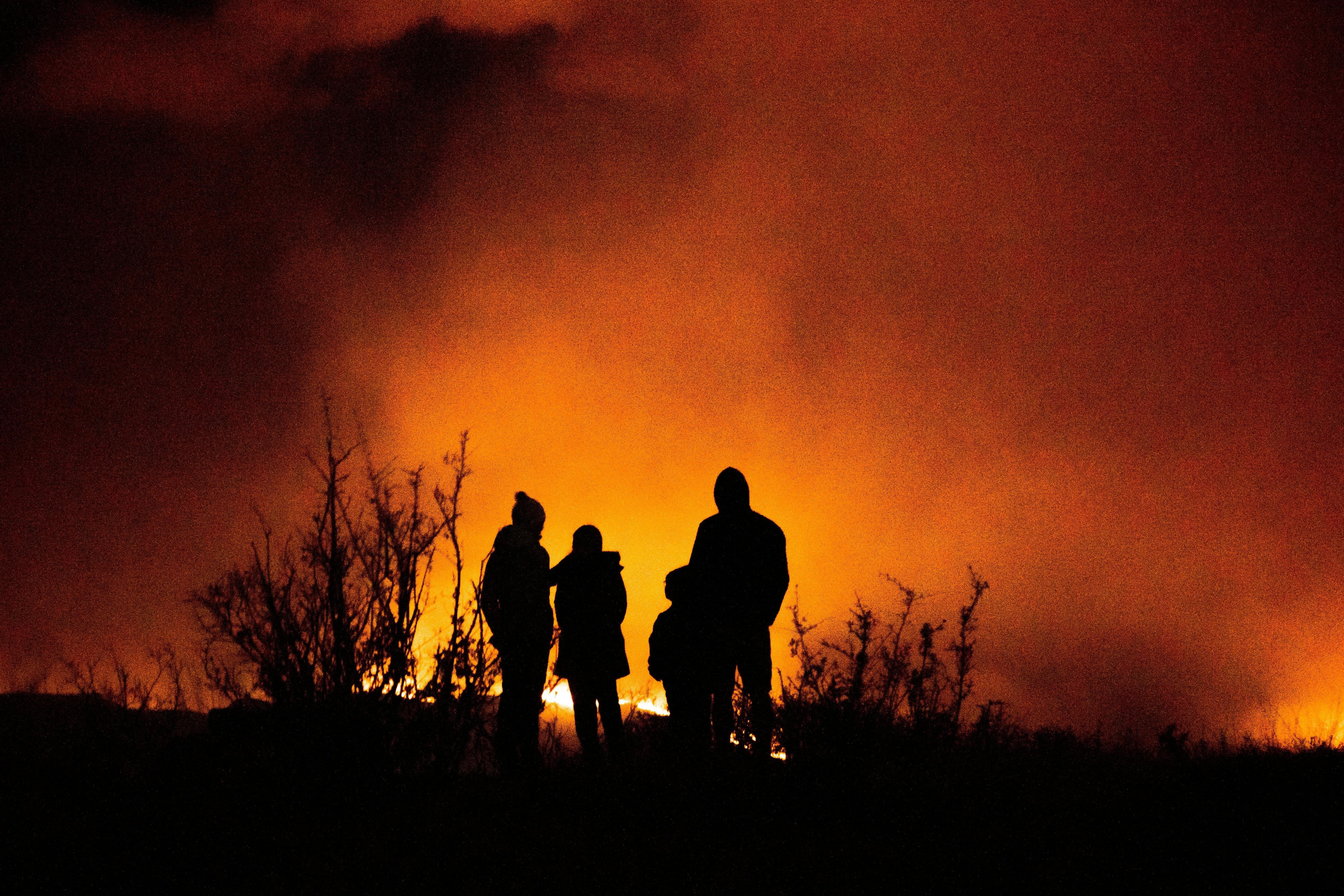 People watching a wildfire at night, with silhouettes outlined against intense flames.