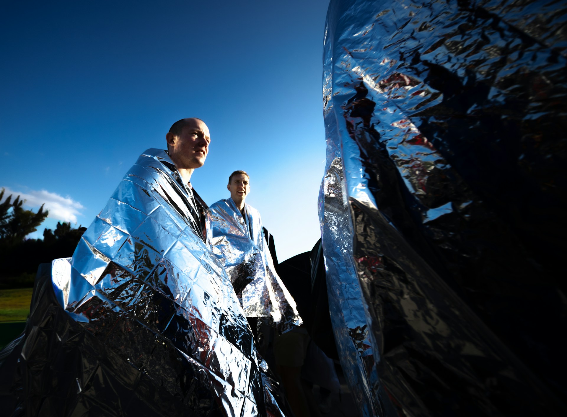 Personas utilizando mantas t&eacute;rmicas de emergencia al aire libre para conservar el calor corporal.