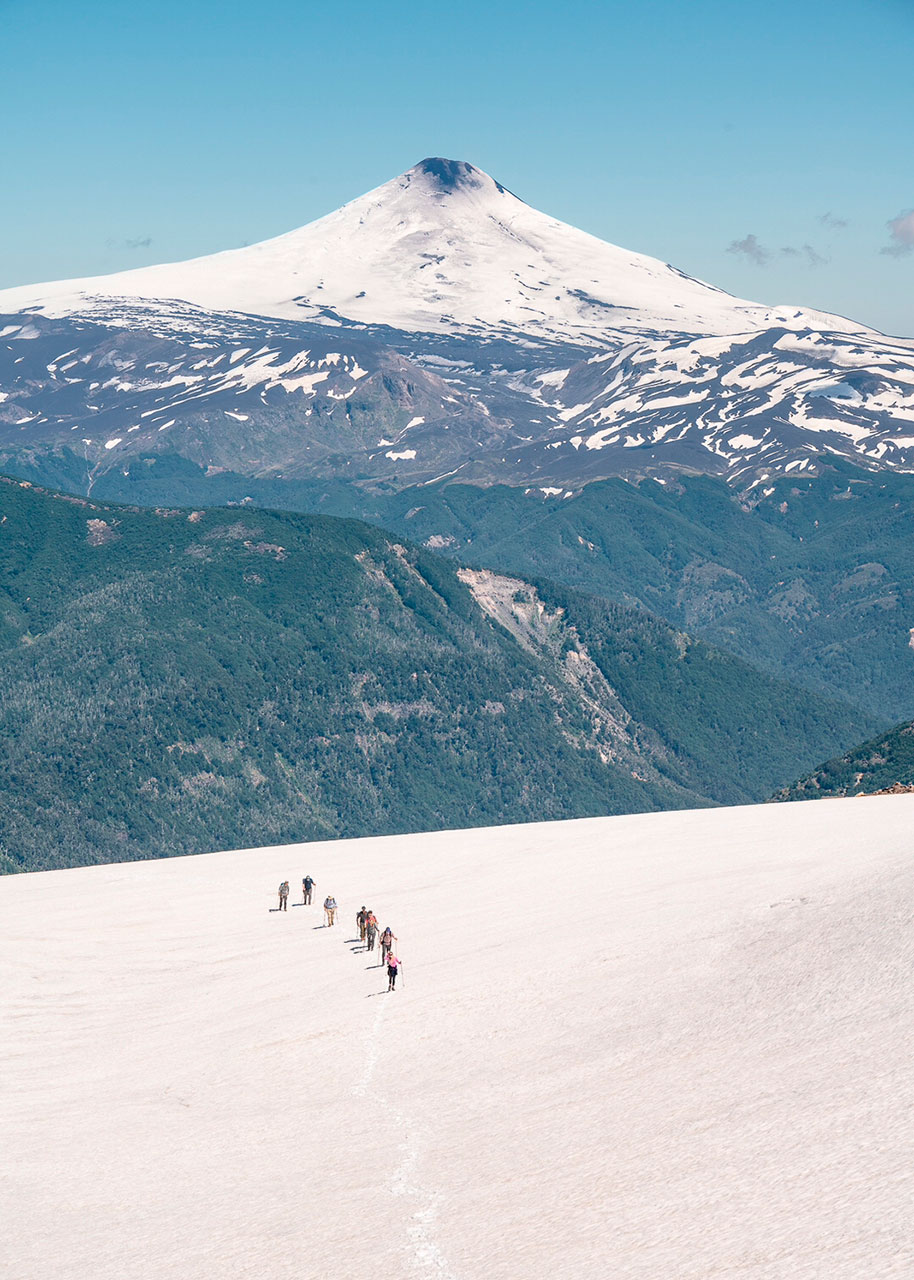 Grupo de montañistas ascendiendo por campo de nieve en el volcán Quetrupillán, con el volcán Villarrica nevado al fondo en la región de La Araucanía, Chile.