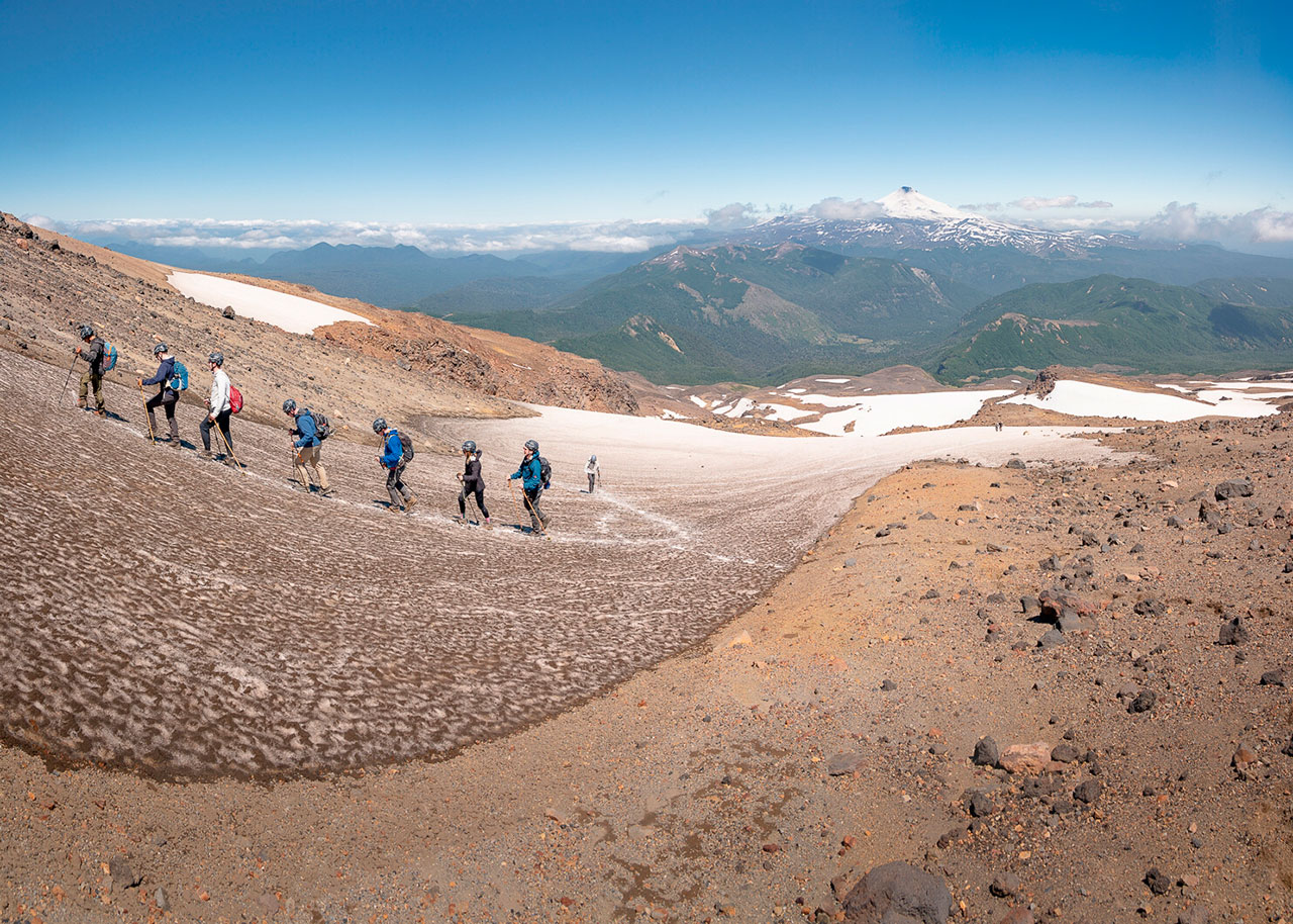 Grupo de excursionistas realizando ascenso al volcán Quetrupillán en Chile, caminando sobre nieve con vista panorámica a la cordillera andina y volcán Villarrica al fondo.