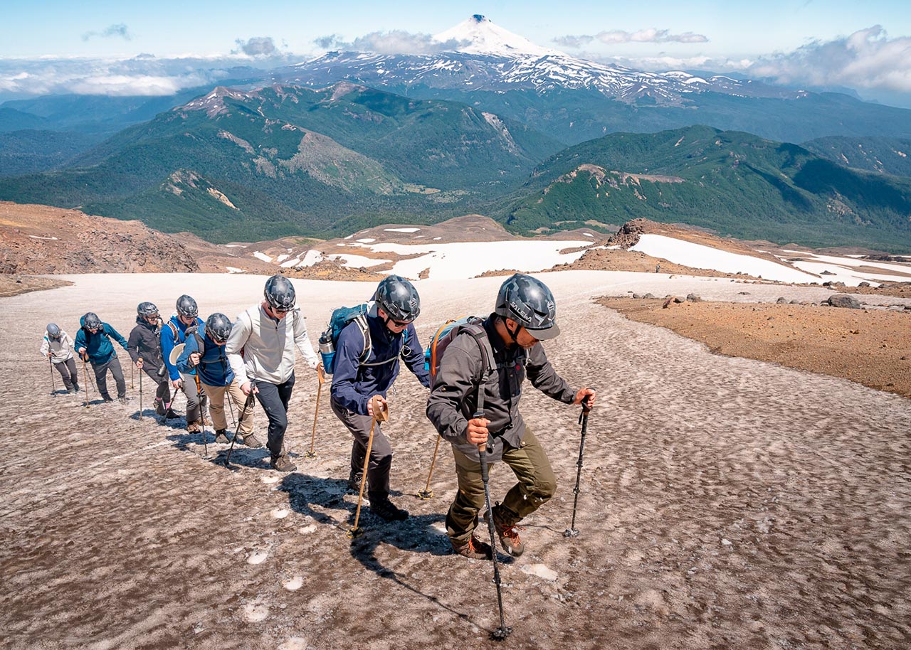 Excursionistas ascendiendo el volcán Quetrupillán con casco, bastones y equipo de montaña sobre nieve, con vista al volcán Villarrica en La Araucanía, Chile.