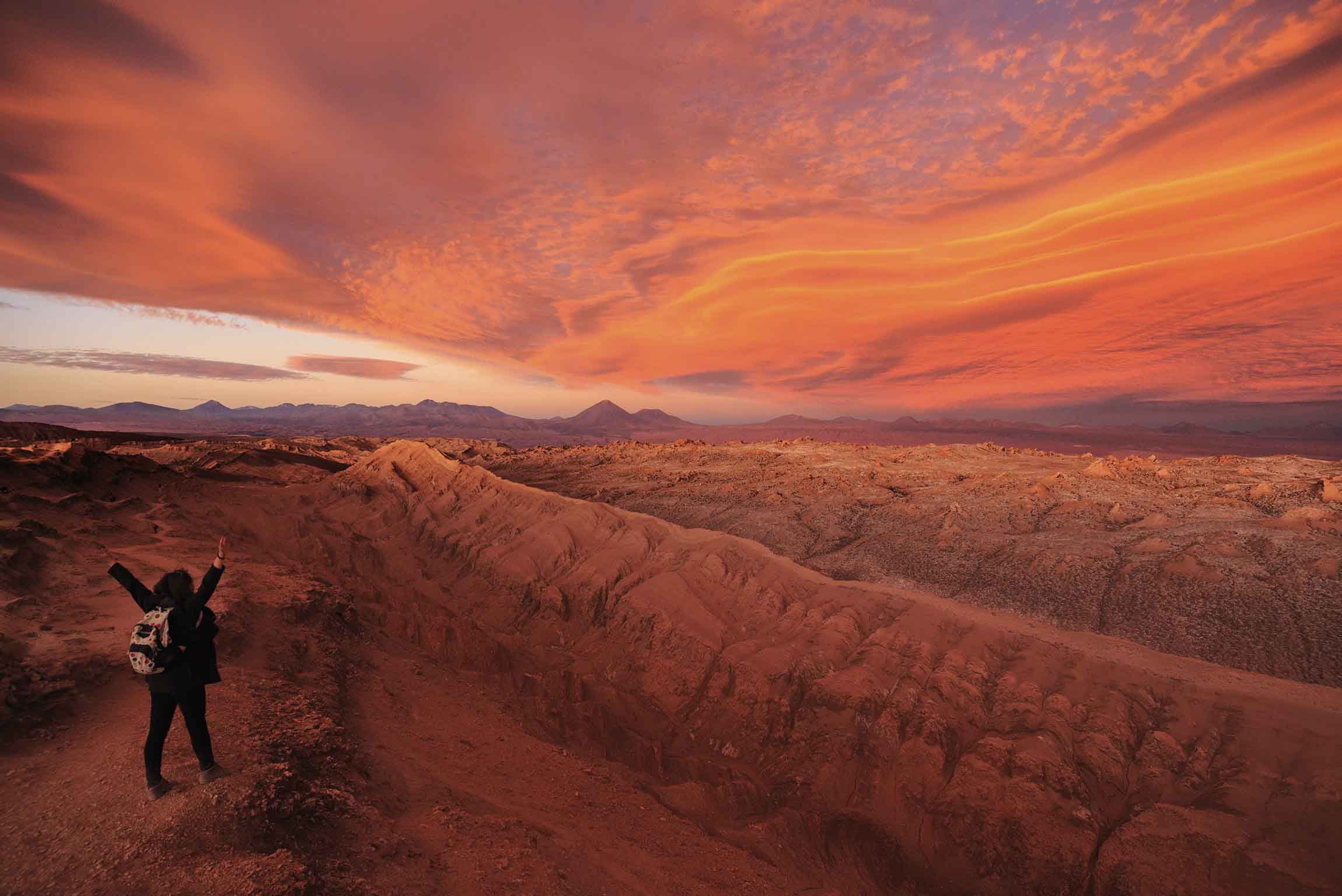 Atardecer anaranjado sobre el Valle de la Luna en San Pedro de Atacama, con formaciones rocosas del desierto y una persona observando el paisaje.