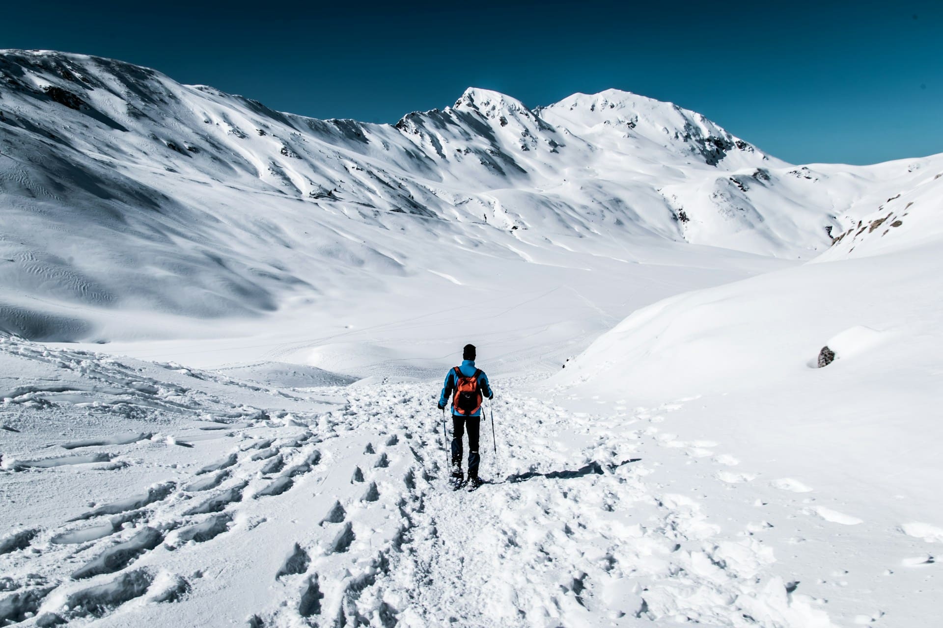 Person walking with trekking poles on a snow-covered path, surrounded by snowy mountains under a clear sky.